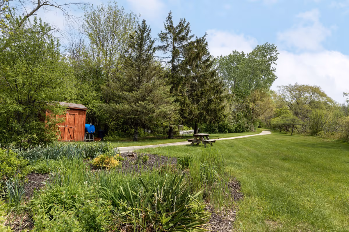 A peaceful outdoor garden area with a variety of green plants and trees. There is a wooden shed on the left side, a picnic table near a paved walking path that curves into the distance, and a clear blue sky with some clouds.