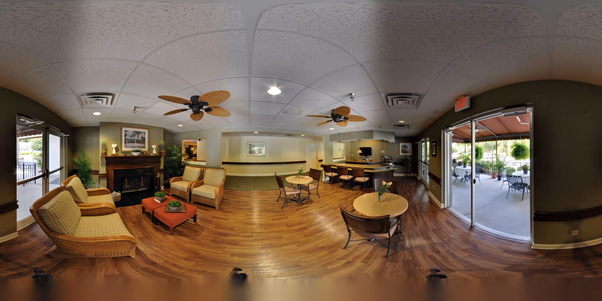 A spacious senior living facility common area with wooden flooring, wicker armchairs with cushions, small round tables with chairs, ceiling fans, and a fireplace. Large glass doors open to an outdoor patio with tables, chairs, and hanging plants.