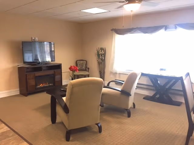 Sunlit common room with two upholstered armchairs facing a TV atop a cabinet with an electric fireplace, a side table, and a window with curtains.