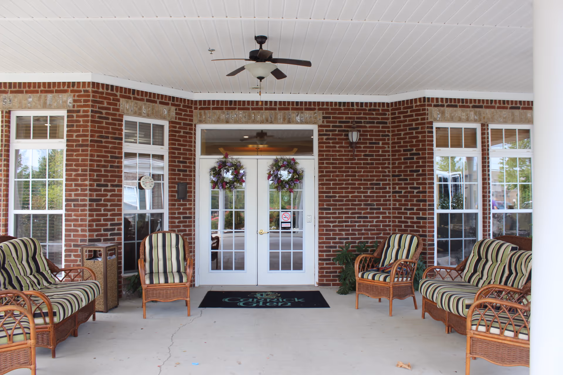 Covered entrance area of Carrick Glen Senior Living with brick walls, white double doors decorated with floral wreaths, and wicker chairs with striped cushions arranged on both sides. A ceiling fan is mounted above, and windows flank the doors.