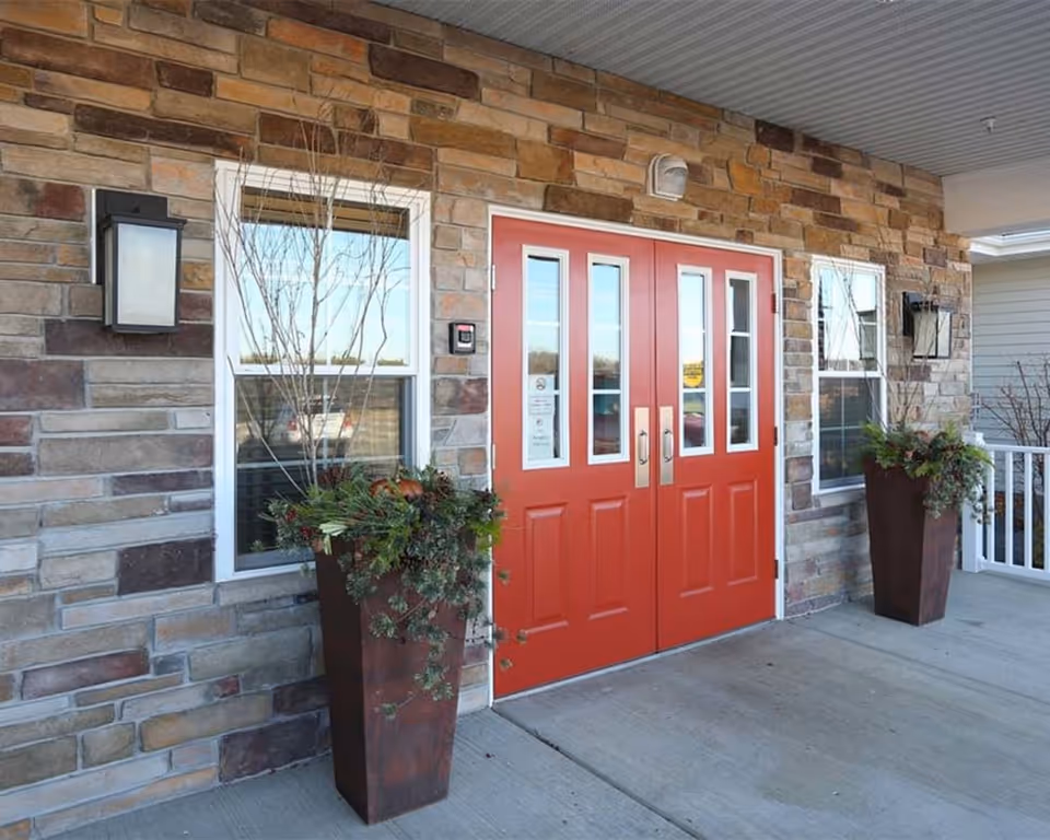 Entrance to a building with double red doors featuring vertical glass panels. The exterior wall is made of multi-colored stone bricks. Two tall planters with greenery and branches flank the doors. Two black wall-mounted lantern-style lights are positioned beside the windows on either side of the doors.