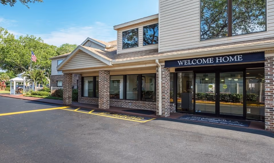 Exterior view of the entrance to a senior living facility with a covered porch supported by brick columns, large windows, and a sign above the entrance that reads 'WELCOME HOME'. There is a paved driveway with yellow no parking markings and an American flag visible in the background near some greenery and a white gazebo.