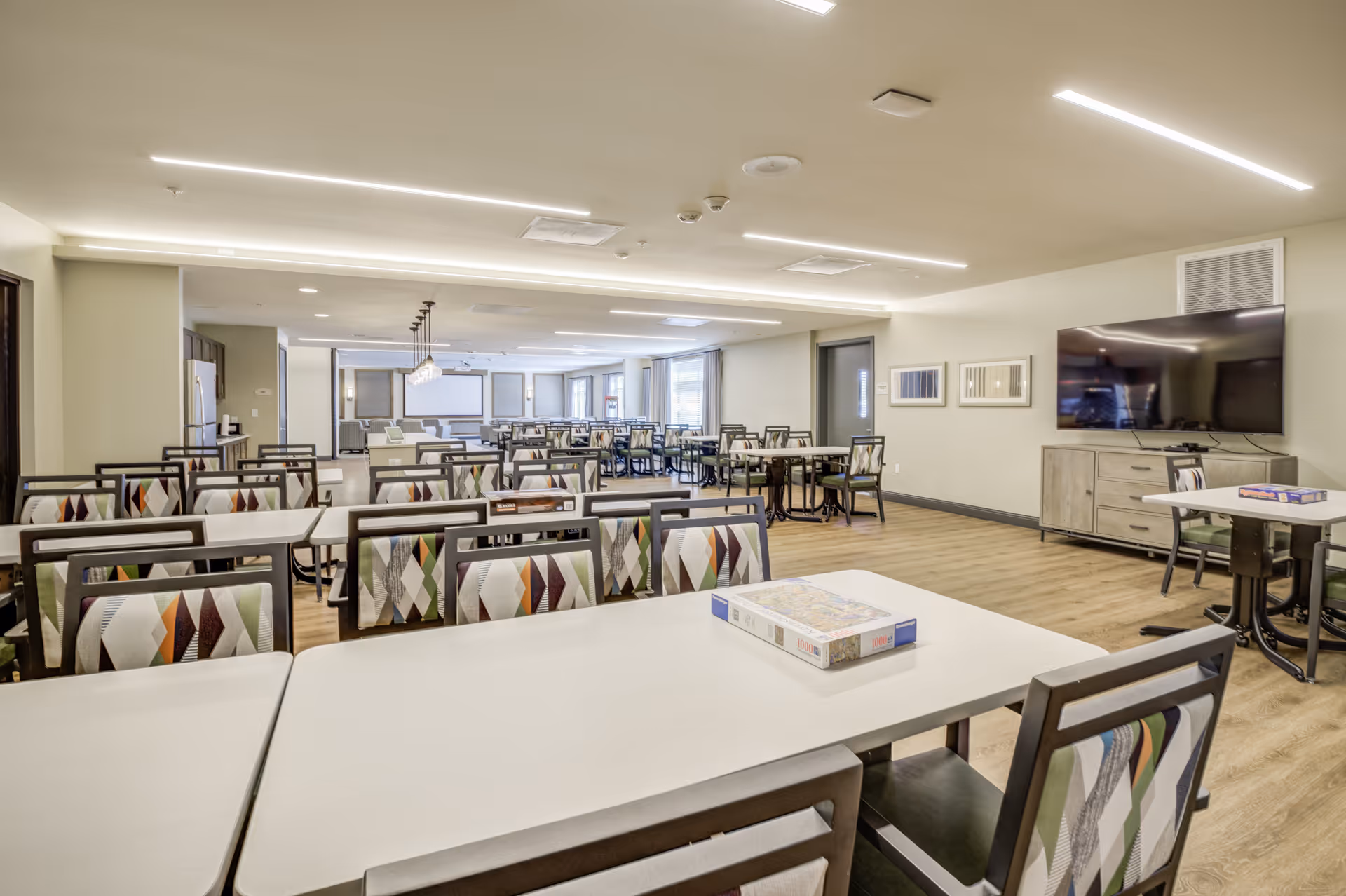 A spacious, well-lit dining room with multiple tables and chairs arranged neatly. The chairs have colorful patterned upholstery. There is a large flat-screen TV mounted on the wall to the right, and a puzzle box is placed on one of the tables in the foreground. The room has light-colored walls, wood flooring, and modern ceiling lighting.