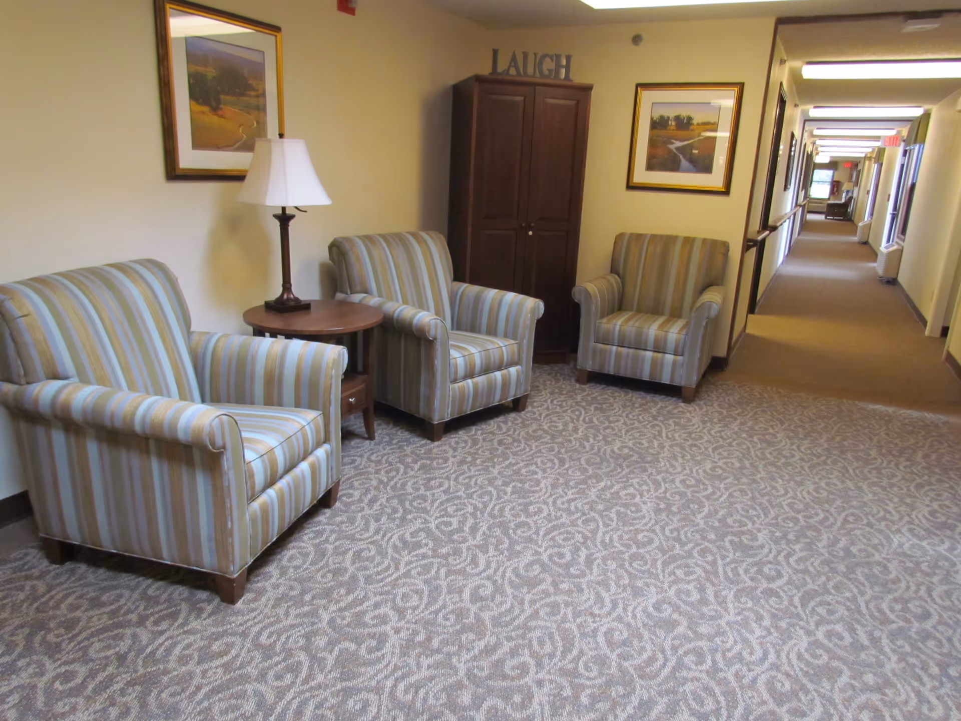 A hallway in Springfield Assisted Living with three striped armchairs arranged around a small round wooden table with a lamp. There are framed landscape paintings on the walls and a wooden cabinet with the word 'LAUGH' on top. The hallway extends into the distance with doors and windows along the right side.