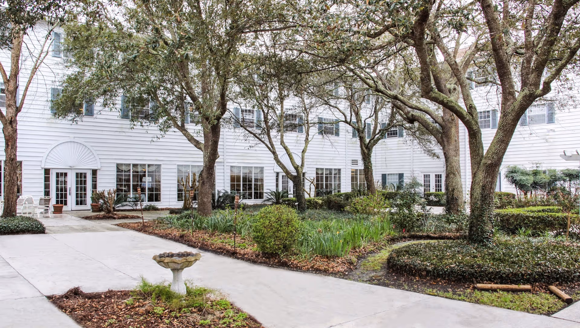 Outdoor courtyard area with trees, shrubs, and a small birdbath in the center. The courtyard is surrounded by a white building with multiple windows and glass doors. There are paved walkways and some outdoor seating visible near the building.