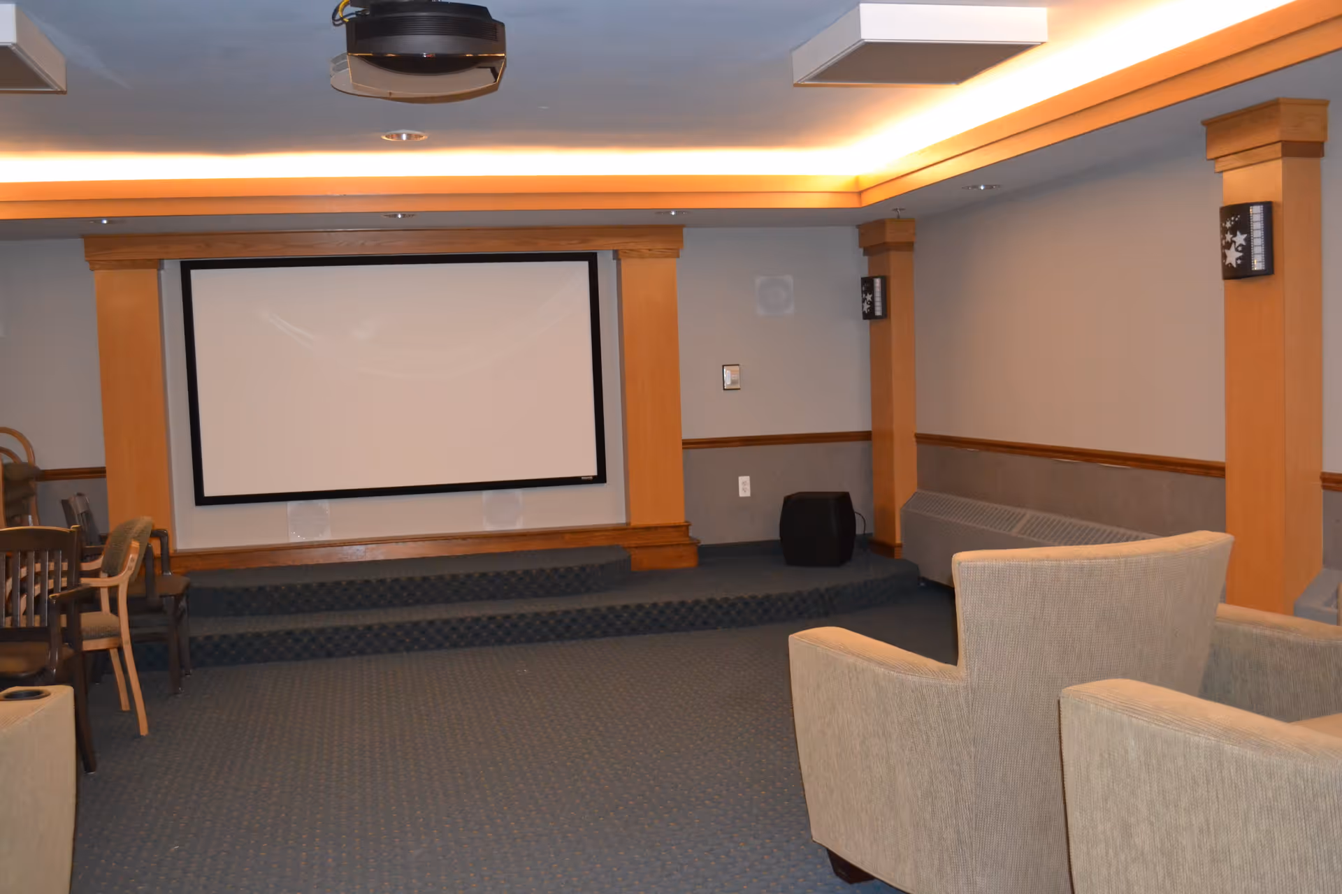 A small theater or media room with a large projection screen mounted on a wooden frame. The room has carpeted flooring, beige walls with wooden trim, and several upholstered chairs arranged facing the screen. There is a ceiling-mounted projector above the seating area.