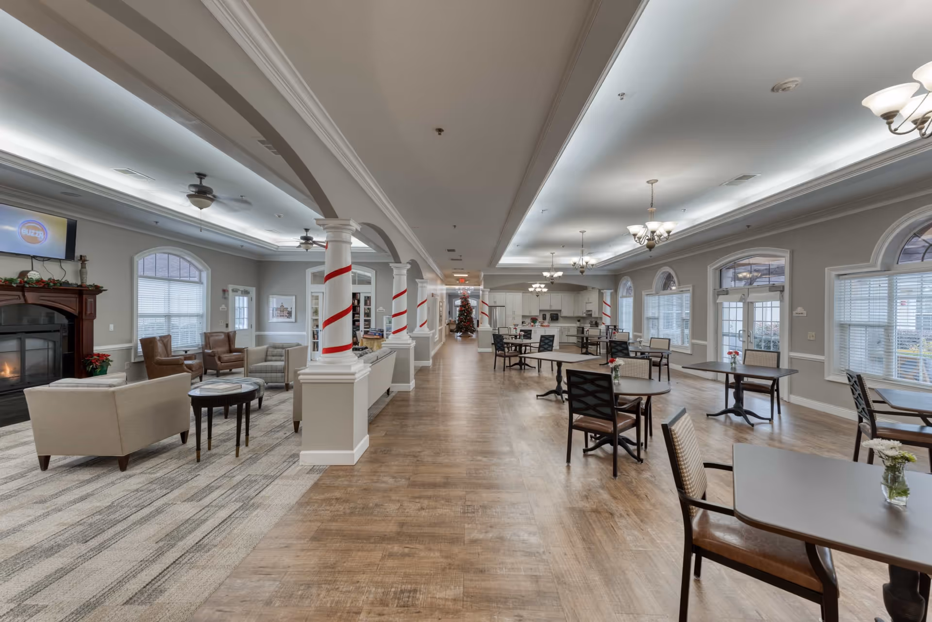 A spacious senior living facility common area with a seating lounge on the left featuring armchairs and sofas around a fireplace, and a dining area on the right with tables and chairs. The room has large windows, ceiling fans, chandeliers, and decorative columns wrapped with red ribbons. A Christmas tree is visible in the background near the kitchen area.