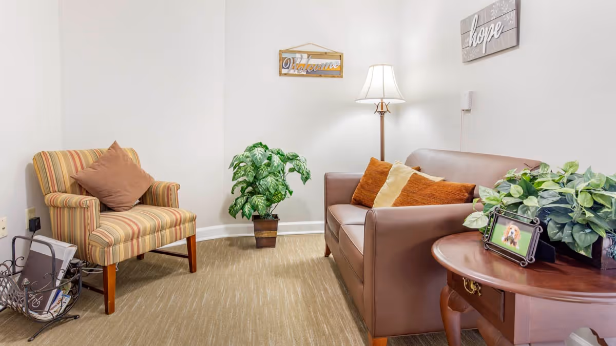 A cozy sitting area with a striped armchair and a brown leather loveseat adorned with orange and beige pillows. A wooden side table holds a framed photo and a potted plant. A floor lamp provides warm lighting, and two wall decorations read 'Welcome' and 'hope'. The room has beige carpeting and white walls.