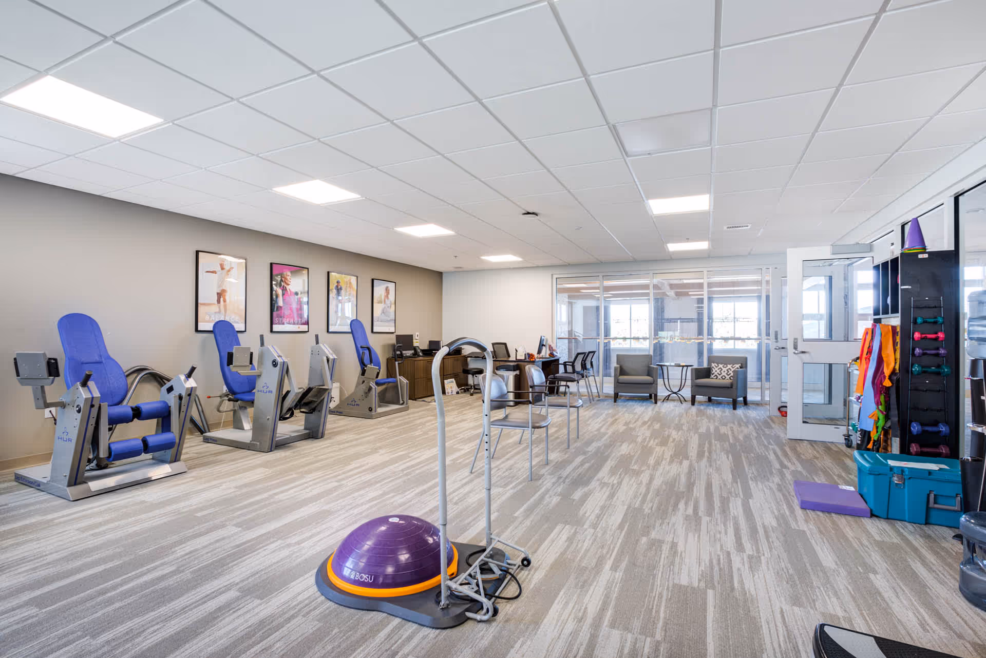 A spacious exercise room with several pieces of fitness equipment including three seated exercise machines with blue padding, a BOSU balance trainer, and various weights and exercise accessories organized on shelves. The room has light-colored carpet flooring, a drop ceiling with recessed lighting, and large windows letting in natural light. There are also a few chairs and tables near the windows, and framed fitness posters on the wall.