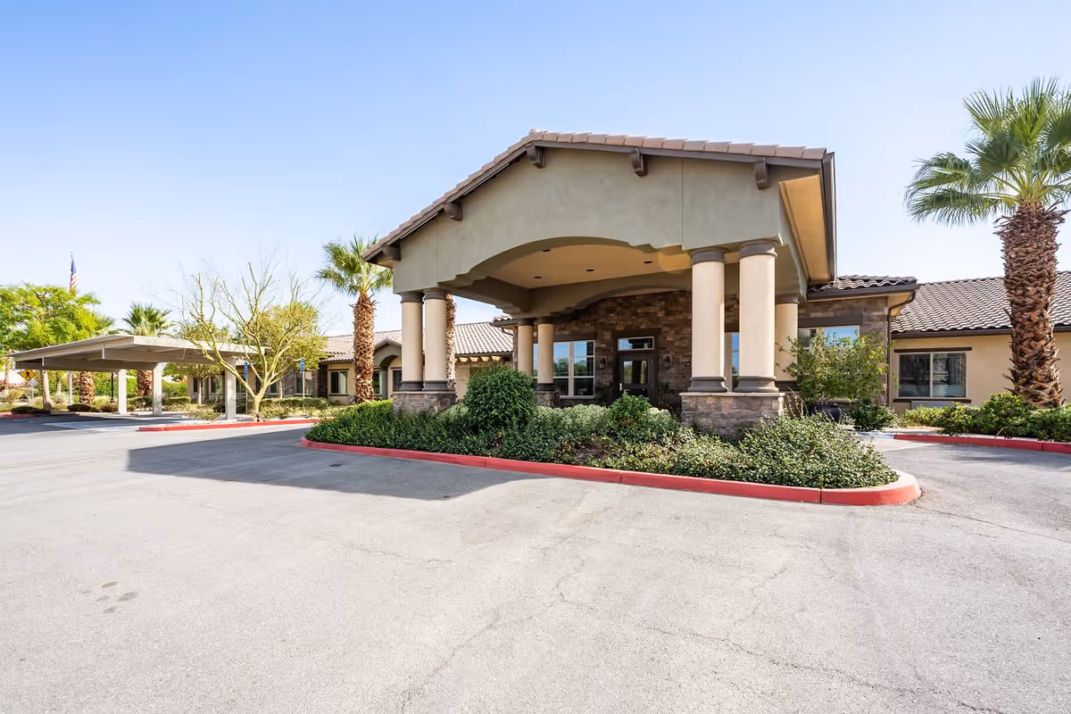 Front exterior view of Caleo Bay Assisted Living and Memory Care facility featuring a covered entrance with columns, surrounding greenery, palm trees, and a clear blue sky.