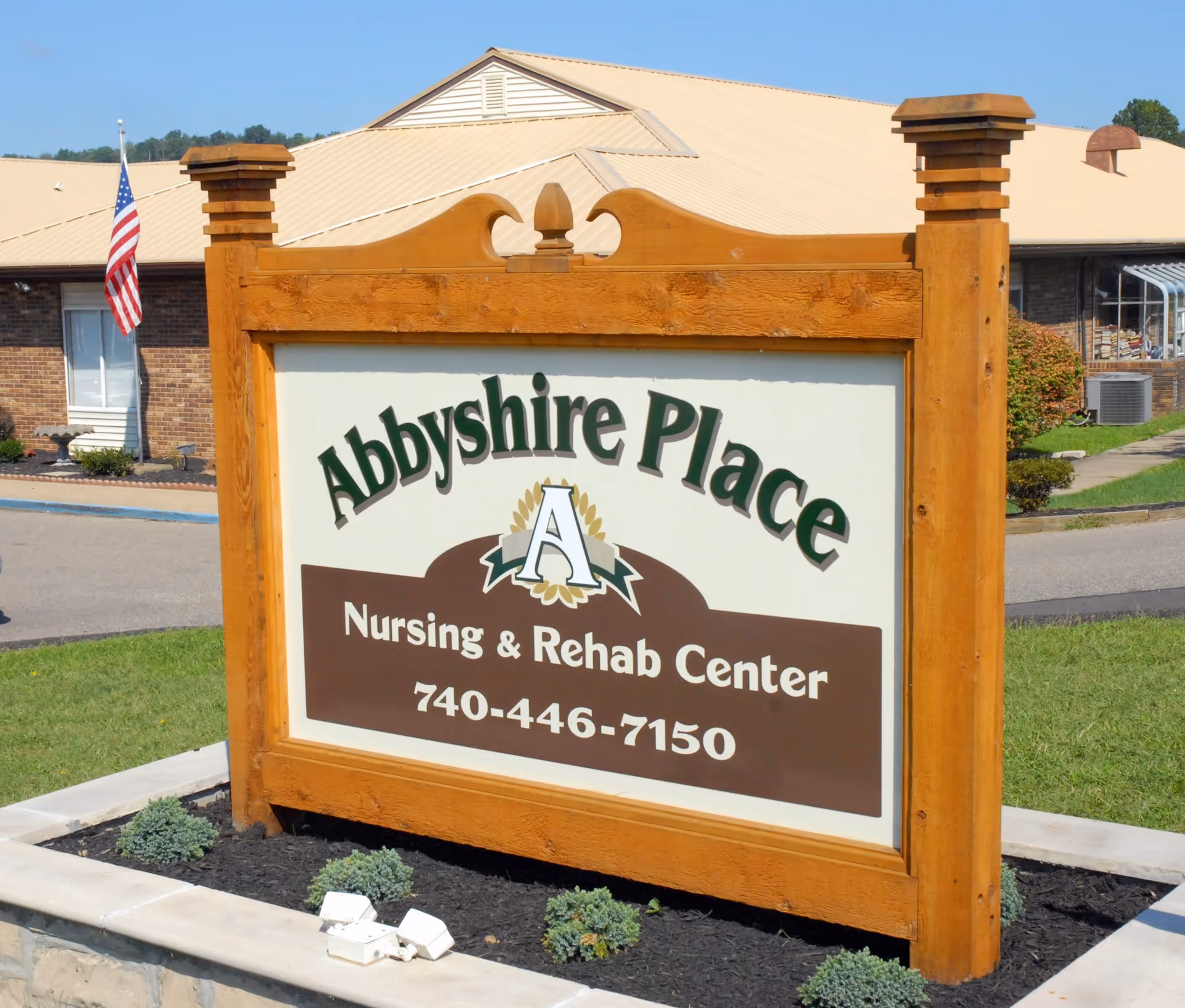 A wooden sign outside a building that reads 'Abbyshire Place Nursing & Rehab Center 740-446-7150' with an American flag and part of a brick building in the background.