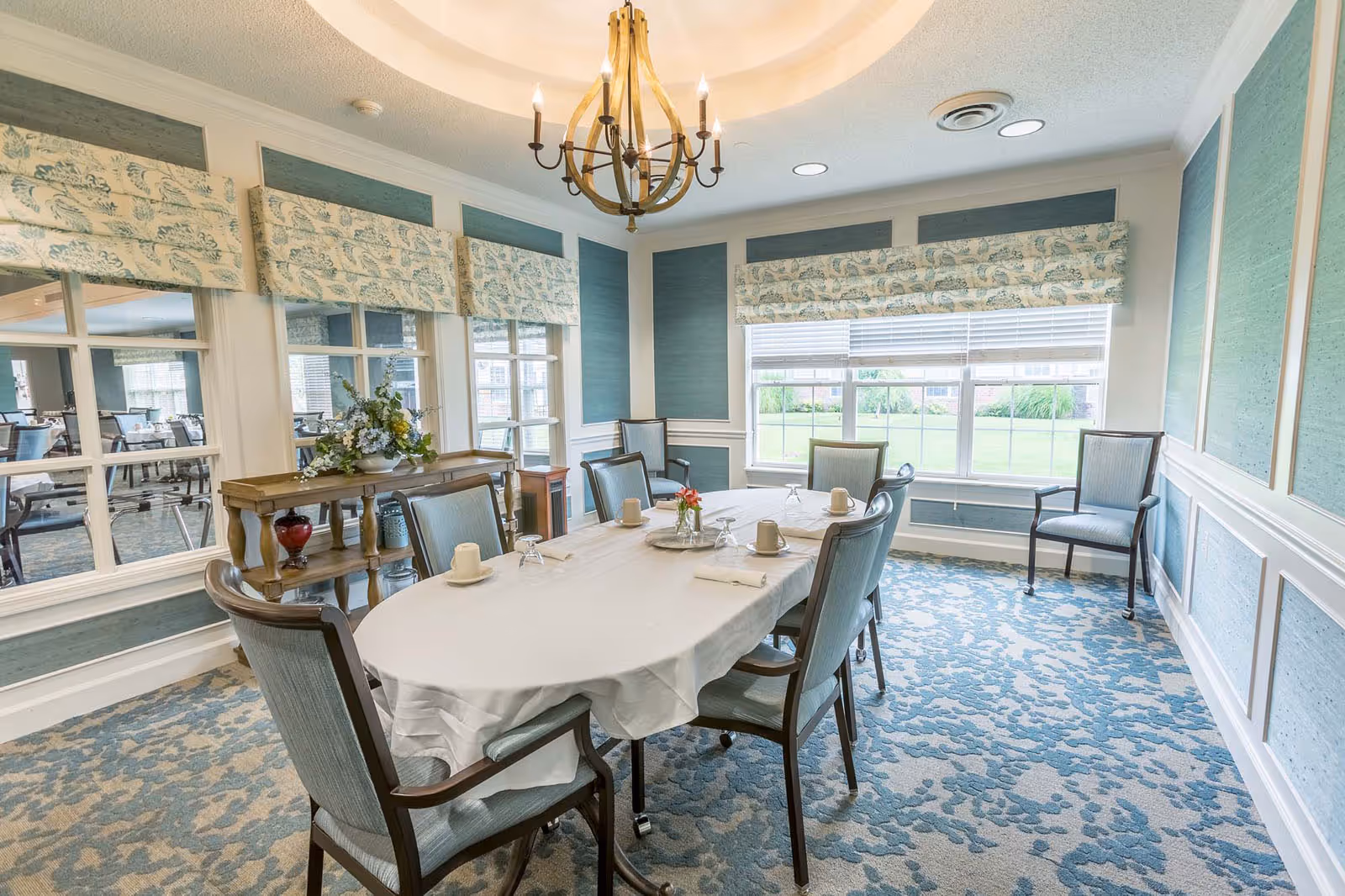 Bright dining room with an oval table set for six, upholstered chairs, a chandelier, and a large window overlooking greenery.
