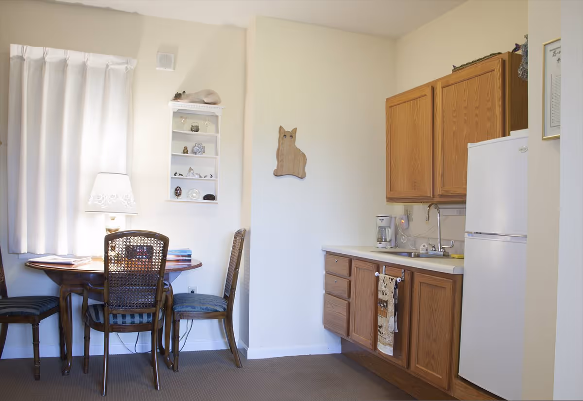 A small kitchen and dining area with wooden cabinets, a white refrigerator, a coffee maker on the counter, and a sink. There is a round wooden table with three chairs, a table lamp, and a window with white curtains. A small wall shelf with decorative items and a wooden cat-shaped wall hanging are also visible.