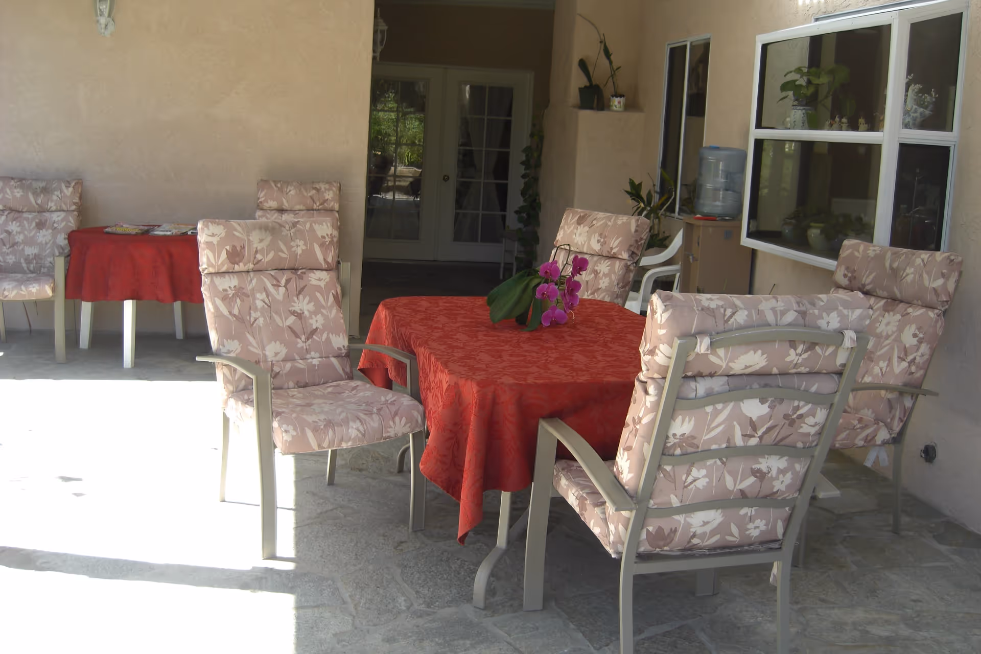 Outdoor patio area with a stone floor, featuring a table covered with a red tablecloth and surrounded by cushioned chairs with floral patterns. There is a purple orchid plant on the table. In the background, there are more chairs and a table with magazines, a water dispenser, potted plants, and a set of glass double doors.
