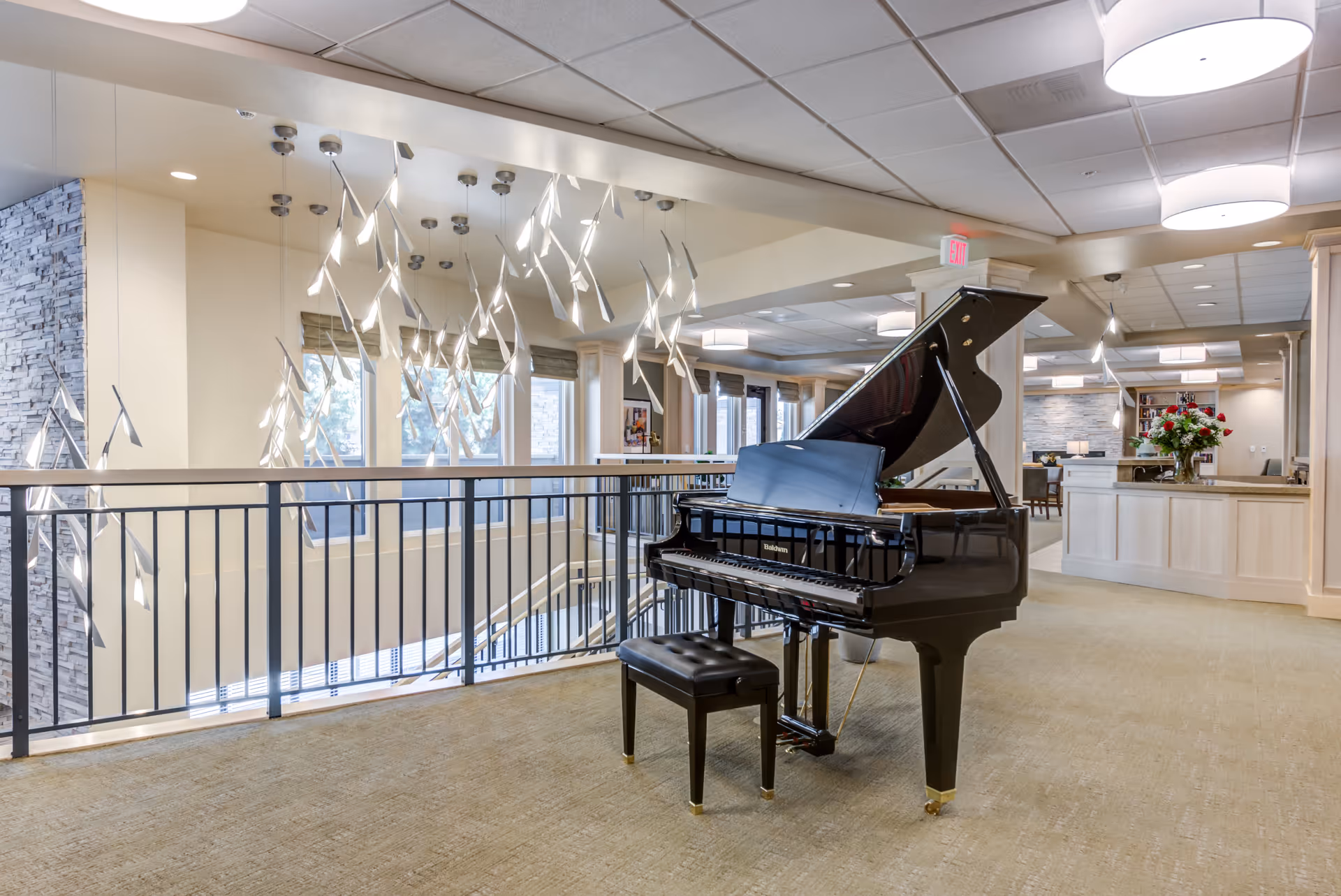 Interior view of a senior living facility featuring a black grand piano with a matching bench on a carpeted floor. The area is well-lit with modern hanging light fixtures and ceiling lights. A railing overlooks a lower level with large windows allowing natural light to enter. In the background, there is a reception or common area with light-colored wood paneling and furniture.