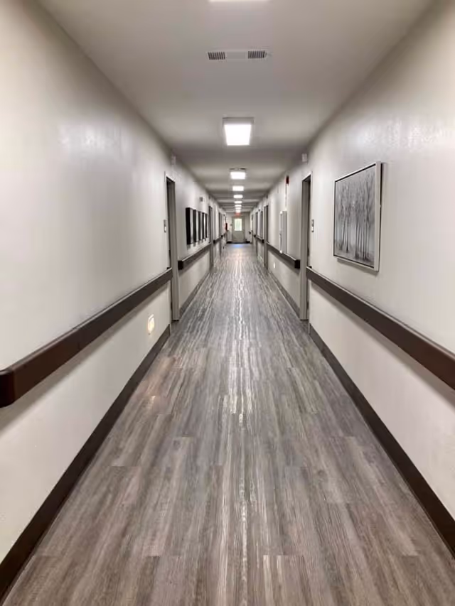 Long, well-lit hallway with light-colored walls and wood-patterned flooring. Handrails run along both sides of the hallway, with several doors and framed pictures on the walls. Ceiling lights are evenly spaced along the corridor.