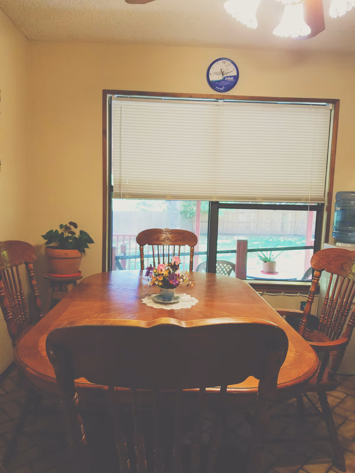 Wooden dining table with chairs and a small flower centerpiece in front of a window with closed blinds.