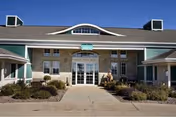 Front entrance of a single-story brick and siding senior living building with a central glass doorway, landscaped planters, and a clear blue sky.