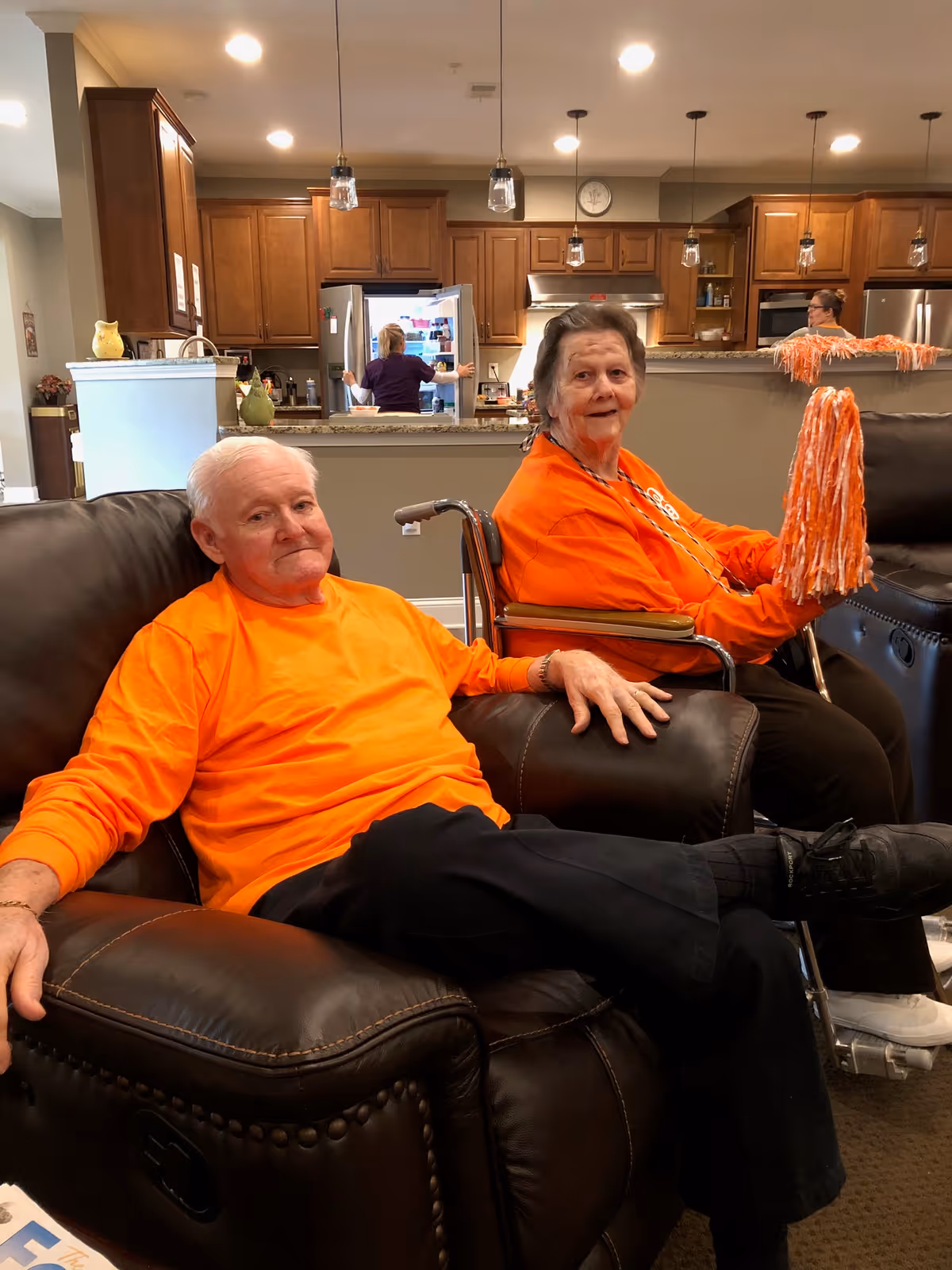 Two elderly individuals wearing bright orange shirts sitting in a living room area. One man is seated in a dark brown leather recliner, and a woman in a wheelchair is holding orange and white pom-poms. In the background, a kitchen with wooden cabinets and two people is visible.