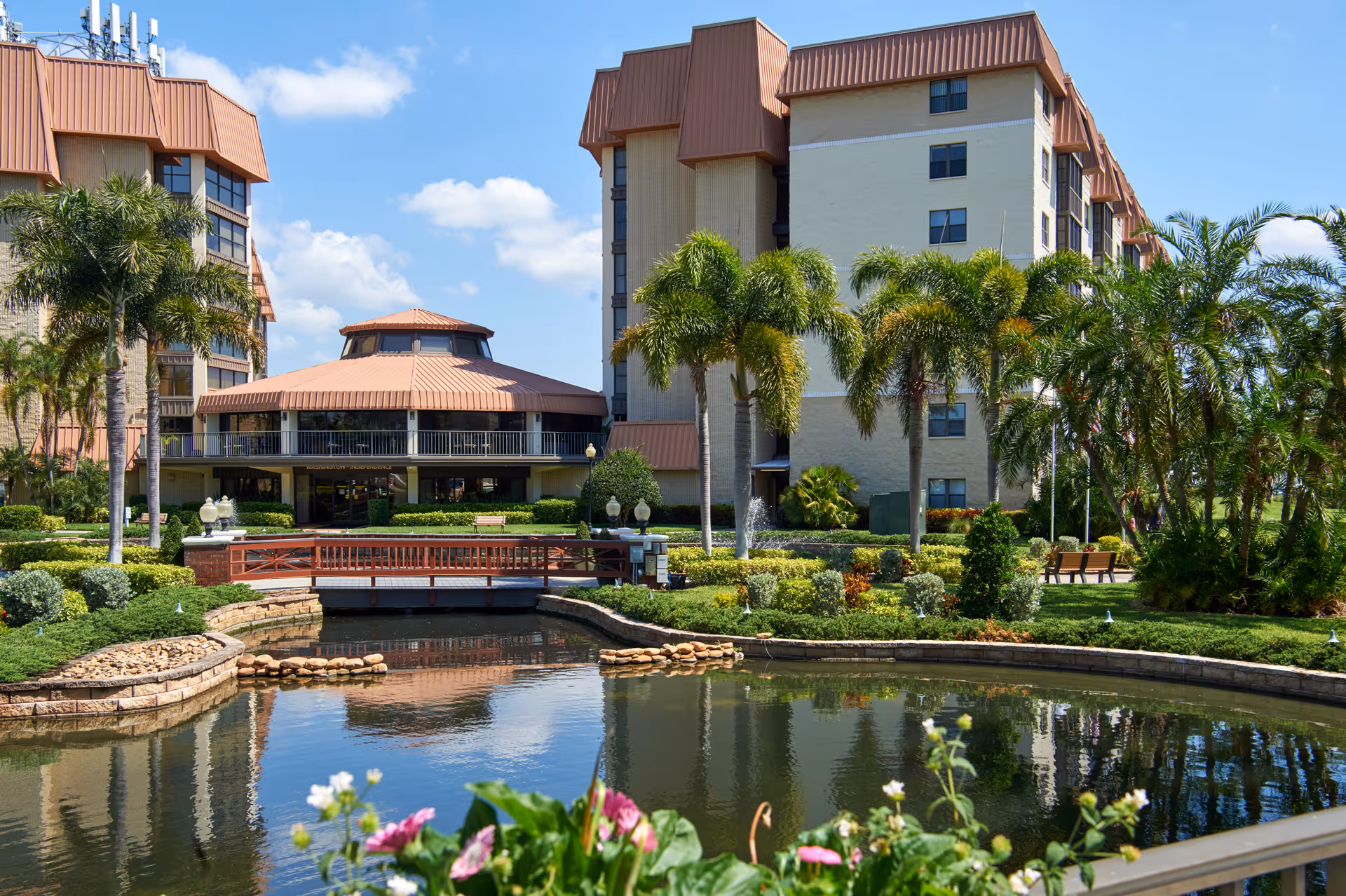 Multi-story senior living building with a central pavilion, landscaped gardens, palm trees and a reflective pond in the foreground.