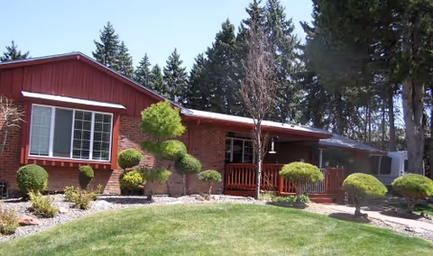 Single-story red brick building front with a manicured lawn, sculpted shrubs, and tall evergreen trees behind it.