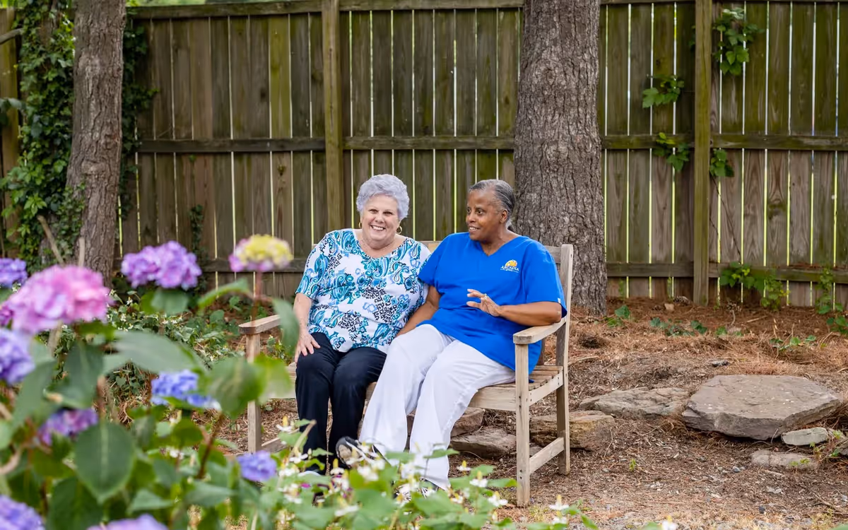 Two women sitting on a wooden bench in a garden area with a wooden fence and trees in the background. One woman is wearing a blue and white patterned shirt and black pants, and the other woman is wearing a blue shirt with the Arcadia logo and white pants. Purple and yellow flowers are visible in the foreground.