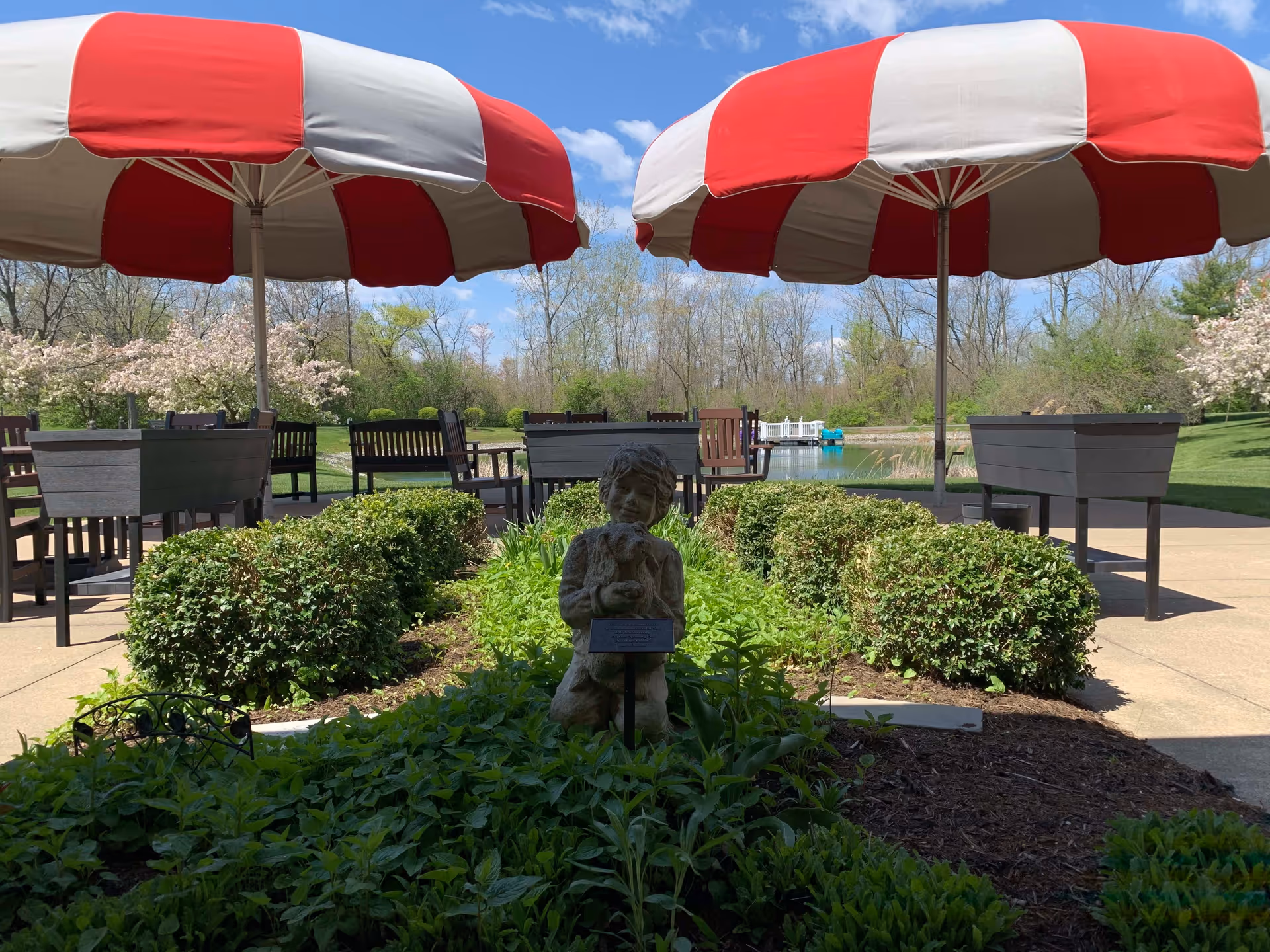 Outdoor patio area with red and white striped umbrellas, wooden benches, and tables surrounded by green bushes and plants. In the foreground, there is a small statue of a child holding a dog. Trees and a pond are visible in the background under a blue sky with some clouds.