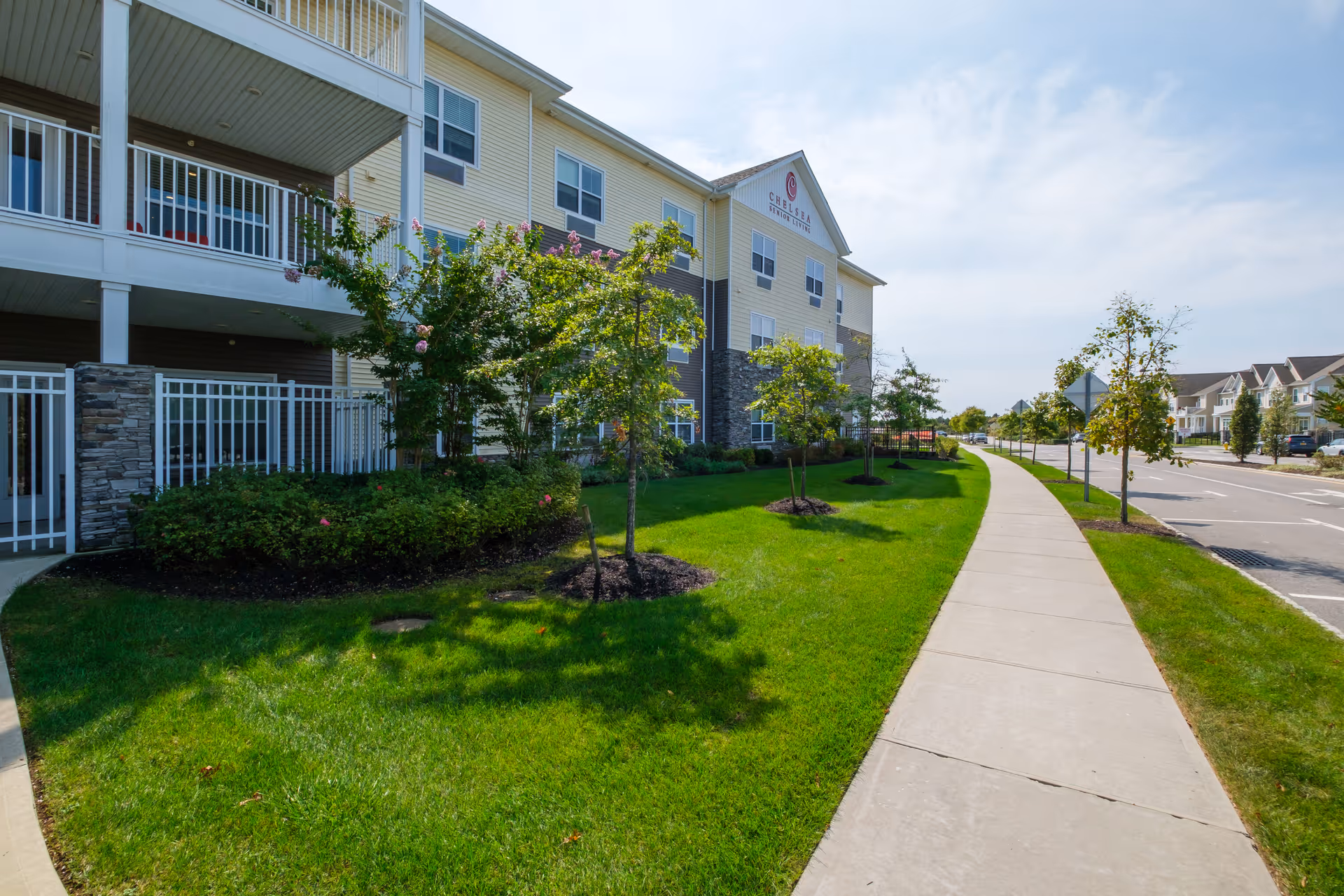 Exterior view of The Chelsea at Brookhaven senior living facility showing a three-story building with balconies, stone and siding facade, well-maintained green lawn, small trees, and a sidewalk running alongside the building and street.