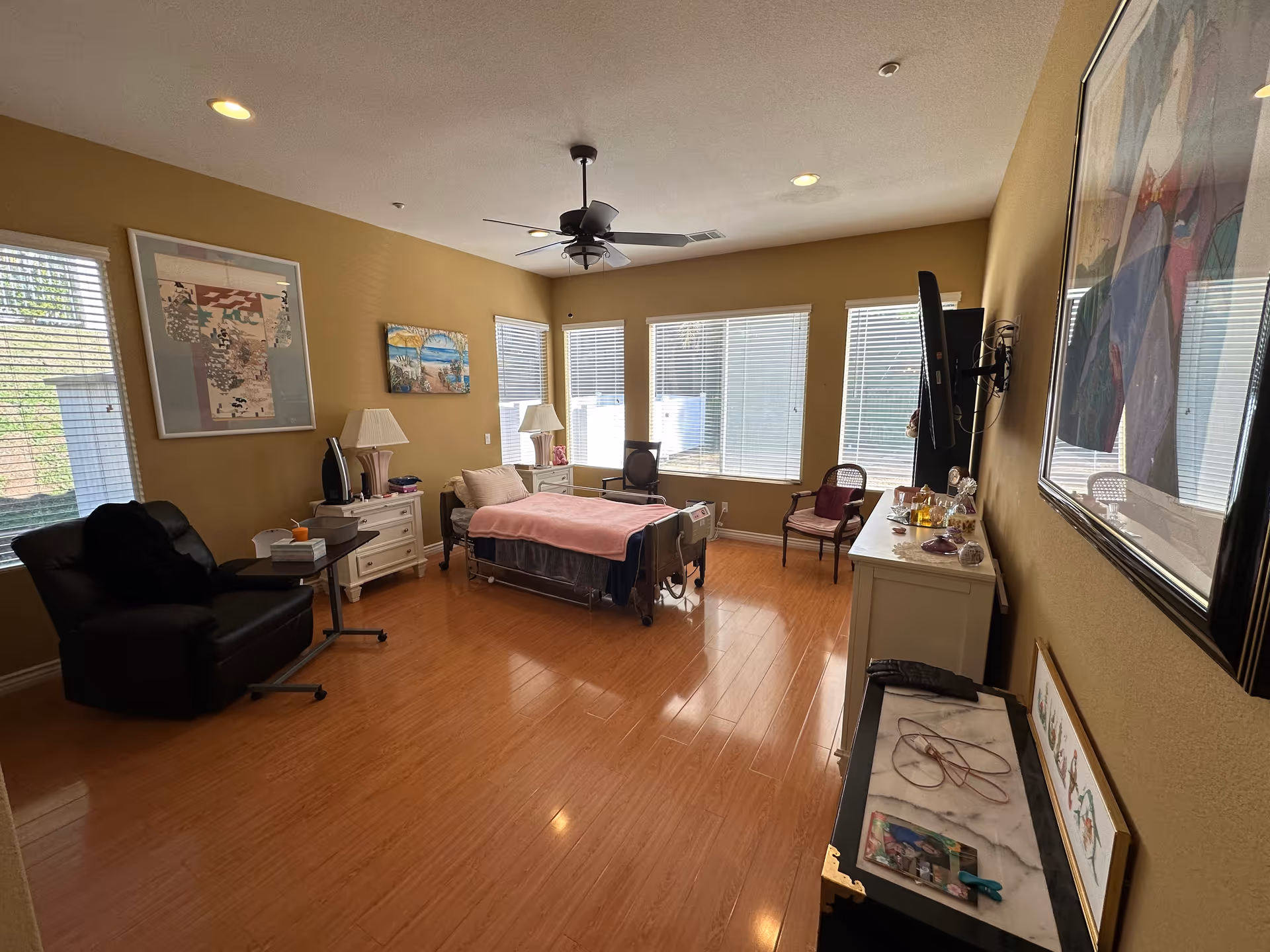 Sunlit senior bedroom with a single bed, recliner, dresser and wall-mounted TV facing large windows.