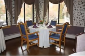 A dining area with a round table covered with a white tablecloth, set with plates, glasses, and napkins. Four wooden chairs with blue cushions surround the table. Large windows with brown curtains provide natural light and a view of trees outside. The walls have floral wallpaper and there is a wooden floor.