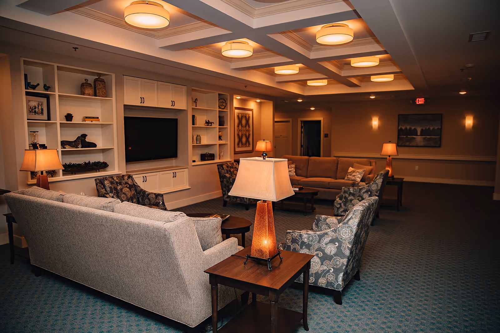 Comfortable senior living lounge with sofas, armchairs, built-in shelving, a TV and warm ambient lighting.