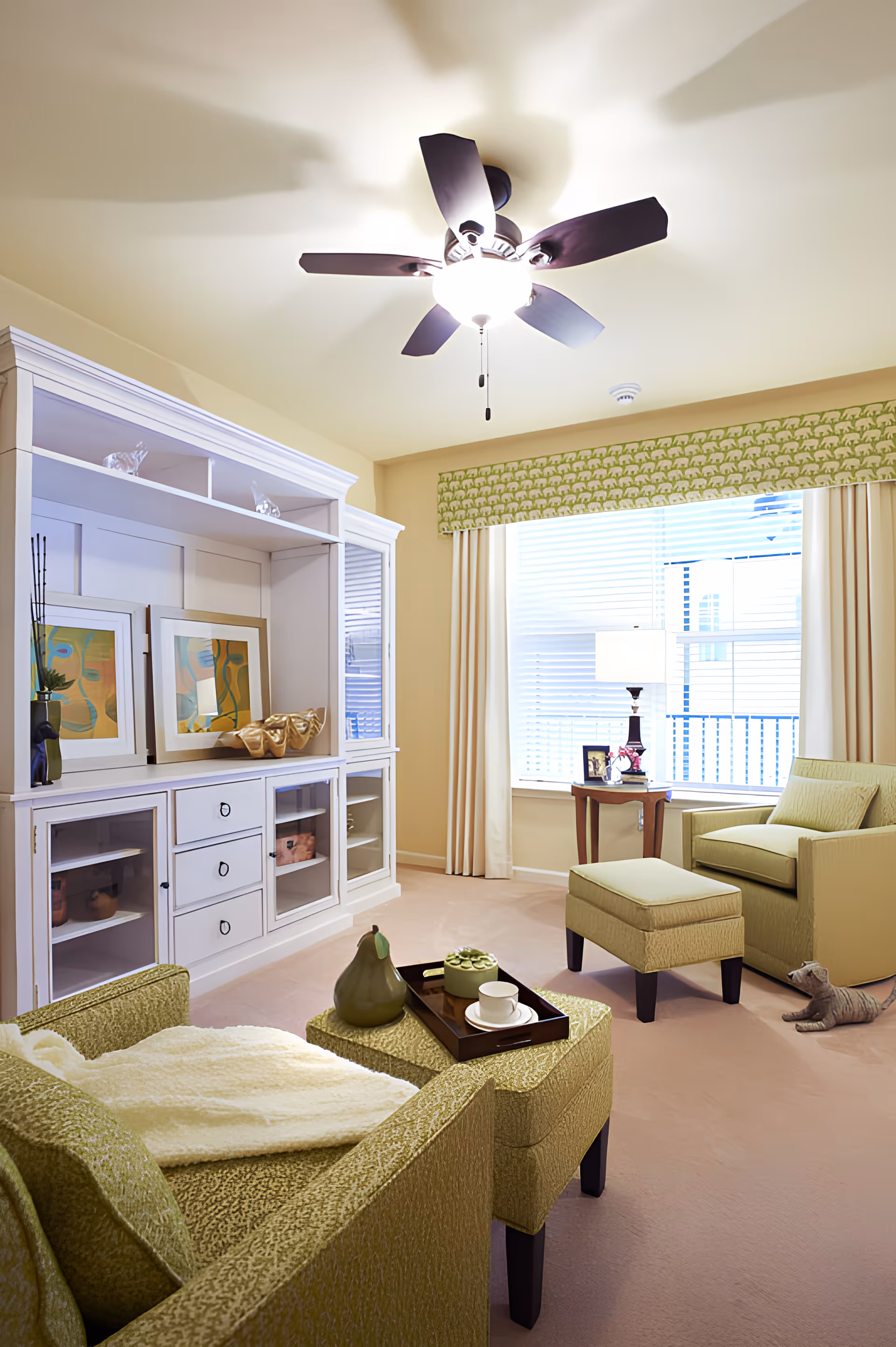 A cozy living room with green upholstered armchairs and ottomans, a white entertainment cabinet with decorative items and framed artwork, a ceiling fan with light, a window with blinds and green patterned valance, a side table with a lamp and picture frame, and a small cat figurine on the floor.