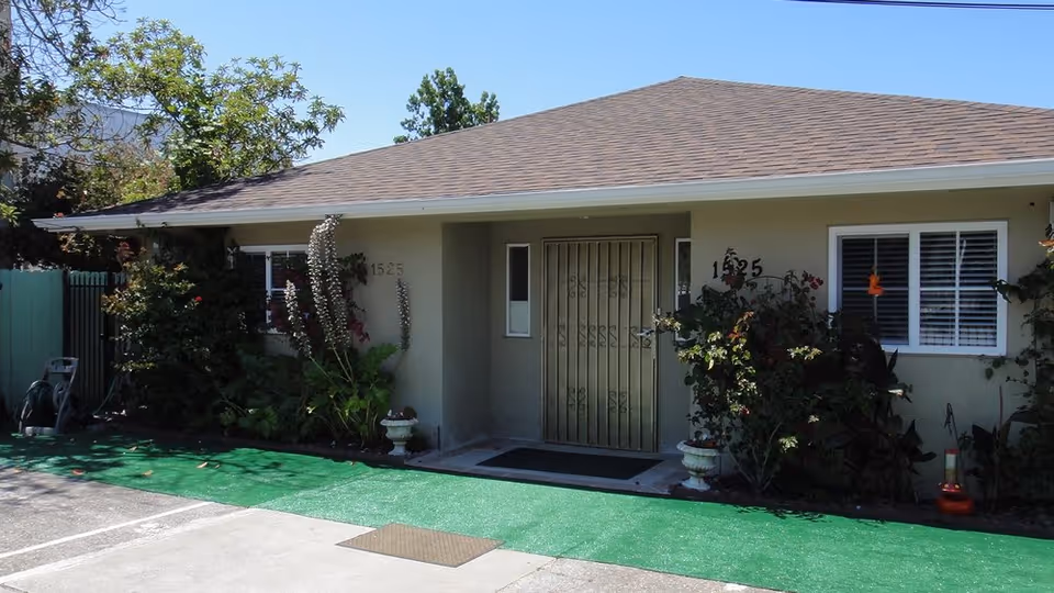 Exterior view of a single-story building with a brown shingled roof and beige walls. The entrance features a metal security door flanked by two small windows. There are plants and shrubs along the front of the building, and the address number 1525 is visible on both sides of the door. The ground in front of the building is covered with green outdoor carpeting and concrete pavement.