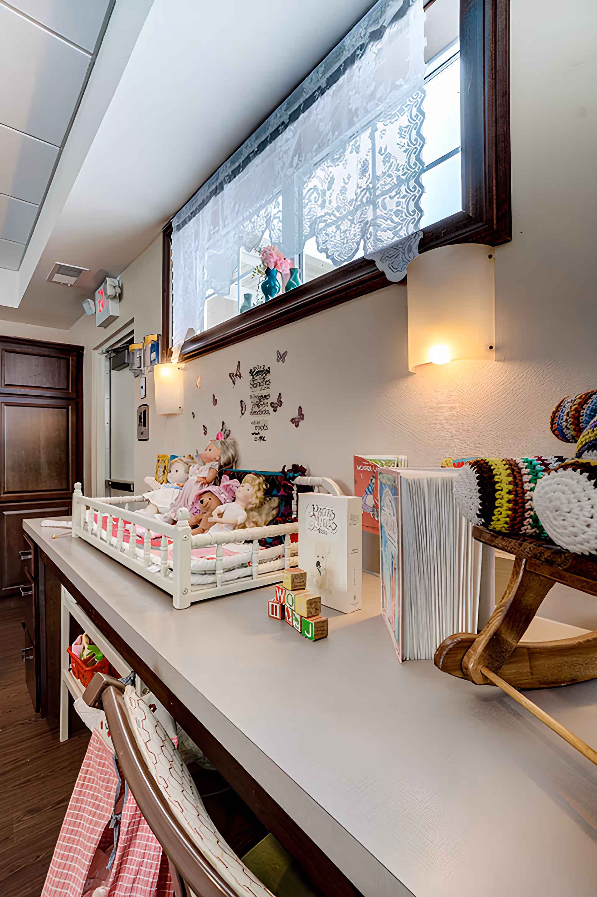 A cozy interior space with a long counter displaying a small white crib filled with dolls, children's books, wooden blocks, and a wooden rocking horse with colorful knitted blankets. The wall above the counter has butterfly decals and two wall-mounted lights. A large window with lace curtains is above the counter, and wooden cabinets and a door are visible in the background.