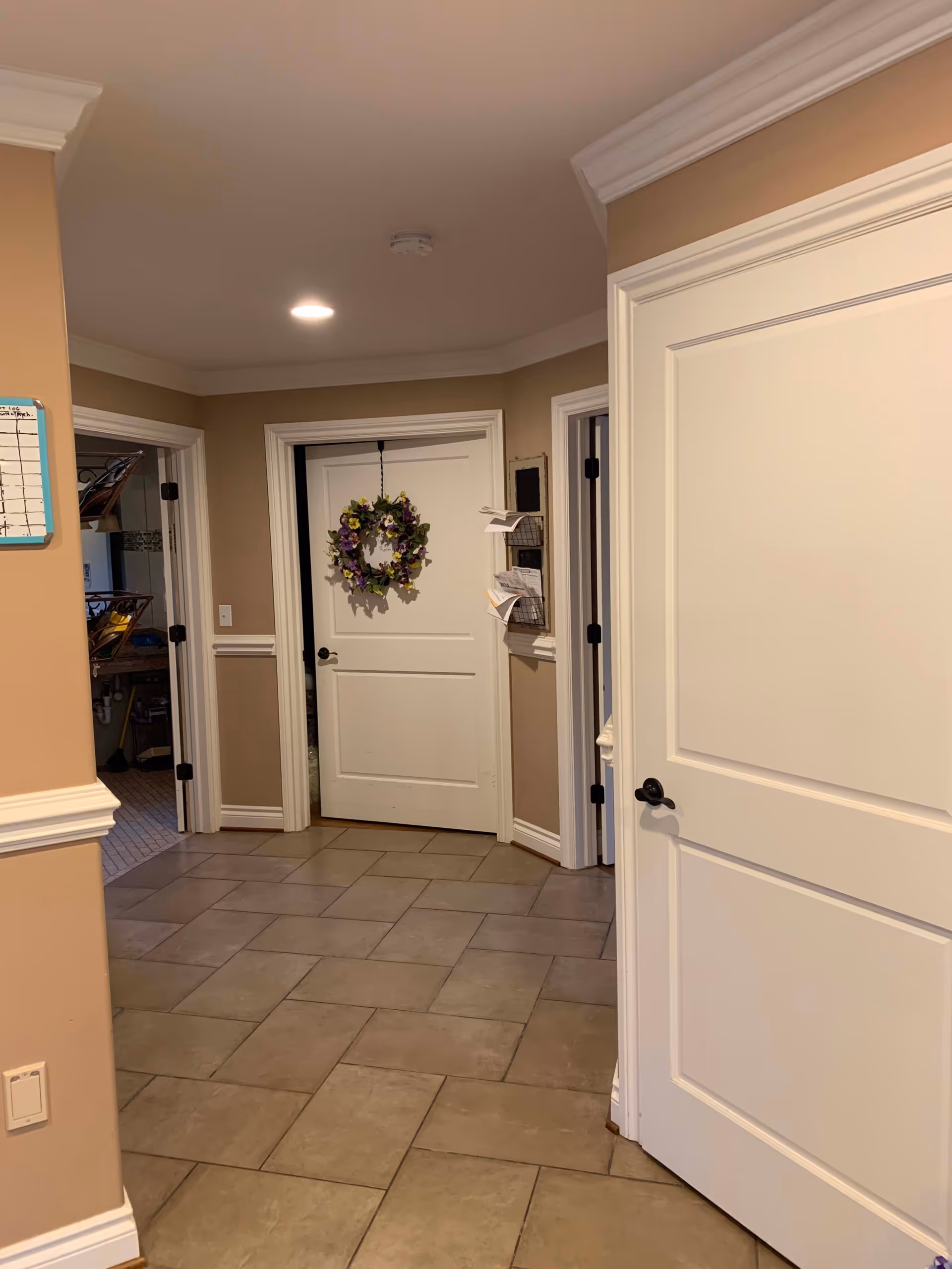 Interior hallway with beige walls and tiled floor, featuring three white doors, one decorated with a purple and yellow floral wreath. There is a wall-mounted organizer with papers and a small whiteboard on the left wall.