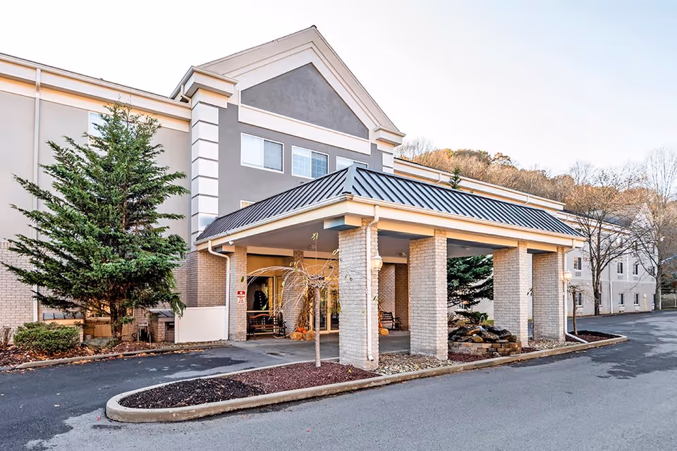 Exterior view of a senior living facility building with a covered entrance supported by brick columns. There are trees and landscaping around the driveway leading to the entrance.