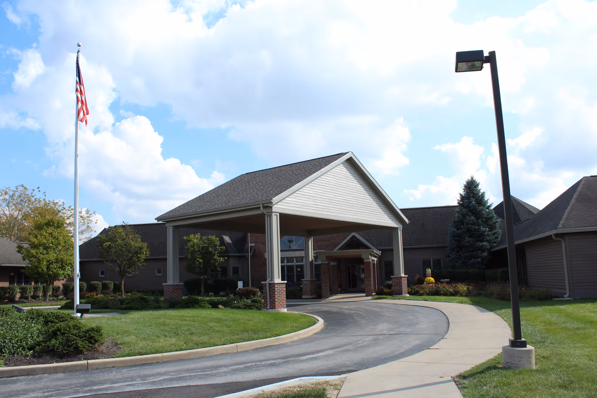 Exterior view of a single-story building with a covered entrance supported by columns. There is a curved driveway leading to the entrance, a sidewalk, a tall flagpole with an American flag, and a street lamp. The building is surrounded by green grass, bushes, and trees under a partly cloudy sky.
