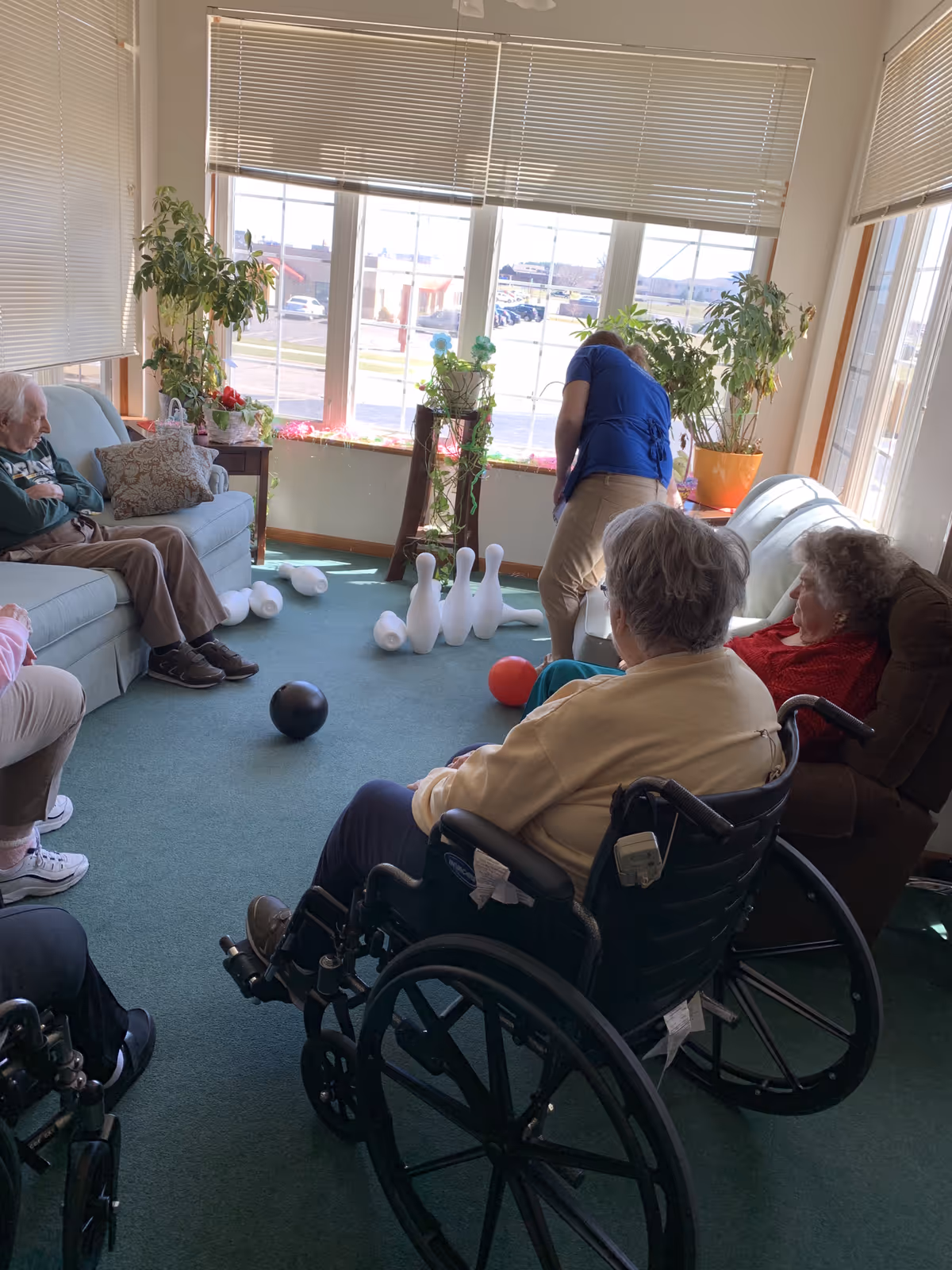 Seniors and a caregiver in a sunlit common room playing indoor bowling with plastic pins and balls.