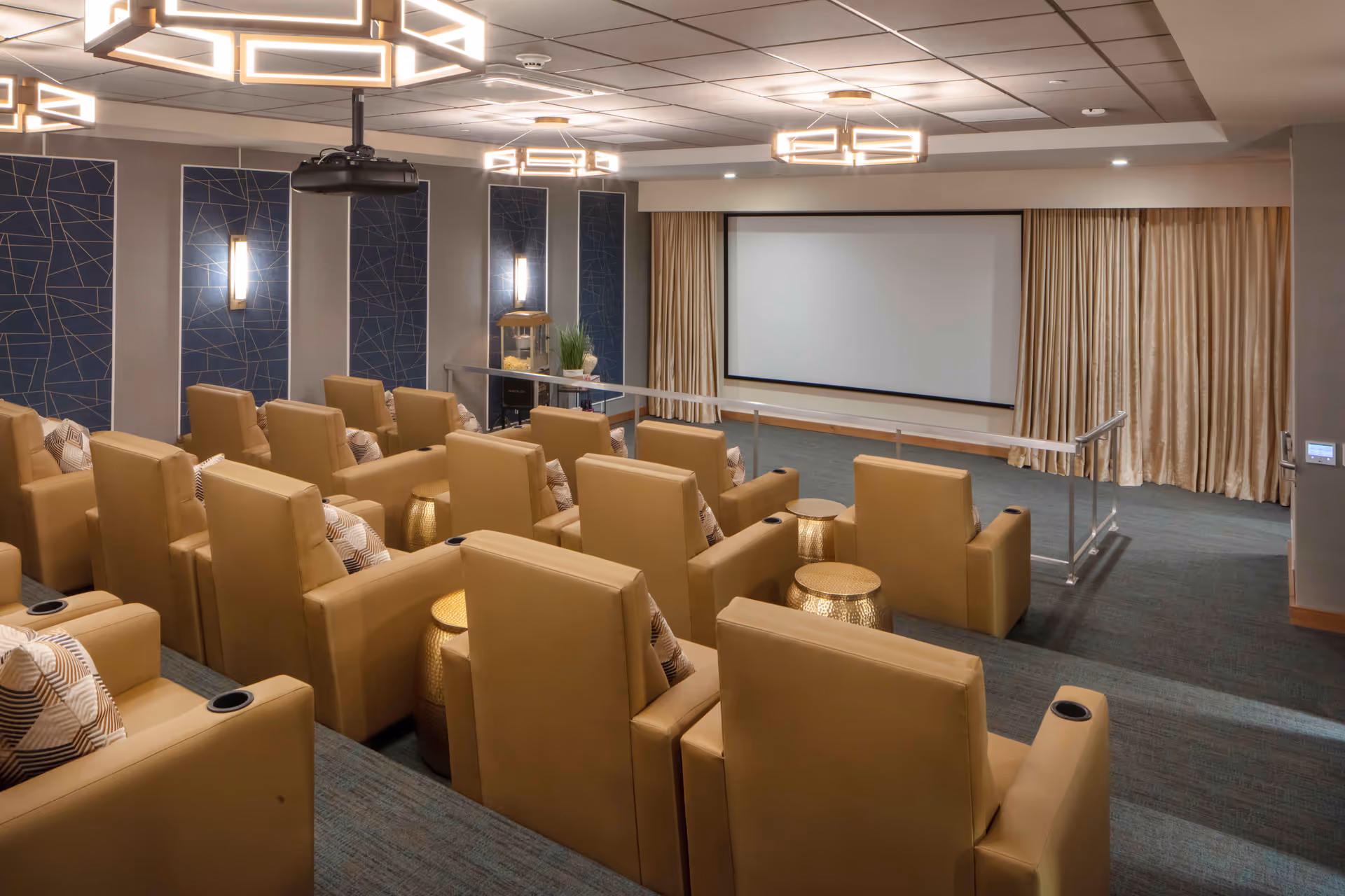 A modern theater room with multiple rows of tan leather recliner chairs, each with a patterned pillow. The room features a large projection screen at the front, blue decorative wall panels with wall sconces, and contemporary ceiling lights. There are small gold side tables between some chairs and a popcorn machine near the front.