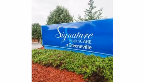 Blue outdoor sign with white text reading 'Signature HealthCARE of Greeneville' surrounded by green shrubs and red mulch, with trees and a cloudy sky in the background.