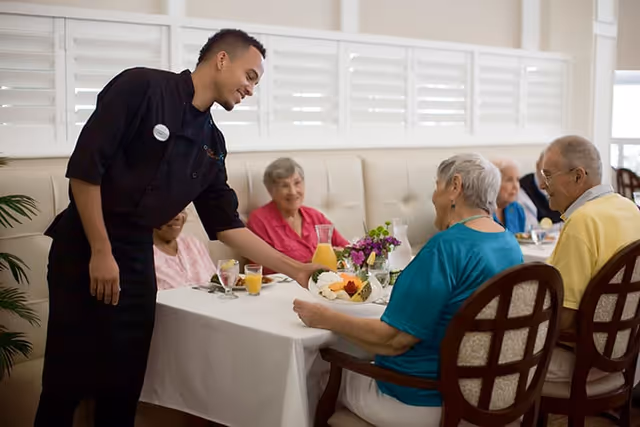 A young waiter serving a plate of food to a group of elderly people seated around a dining table in a bright dining room with white walls and large windows.
