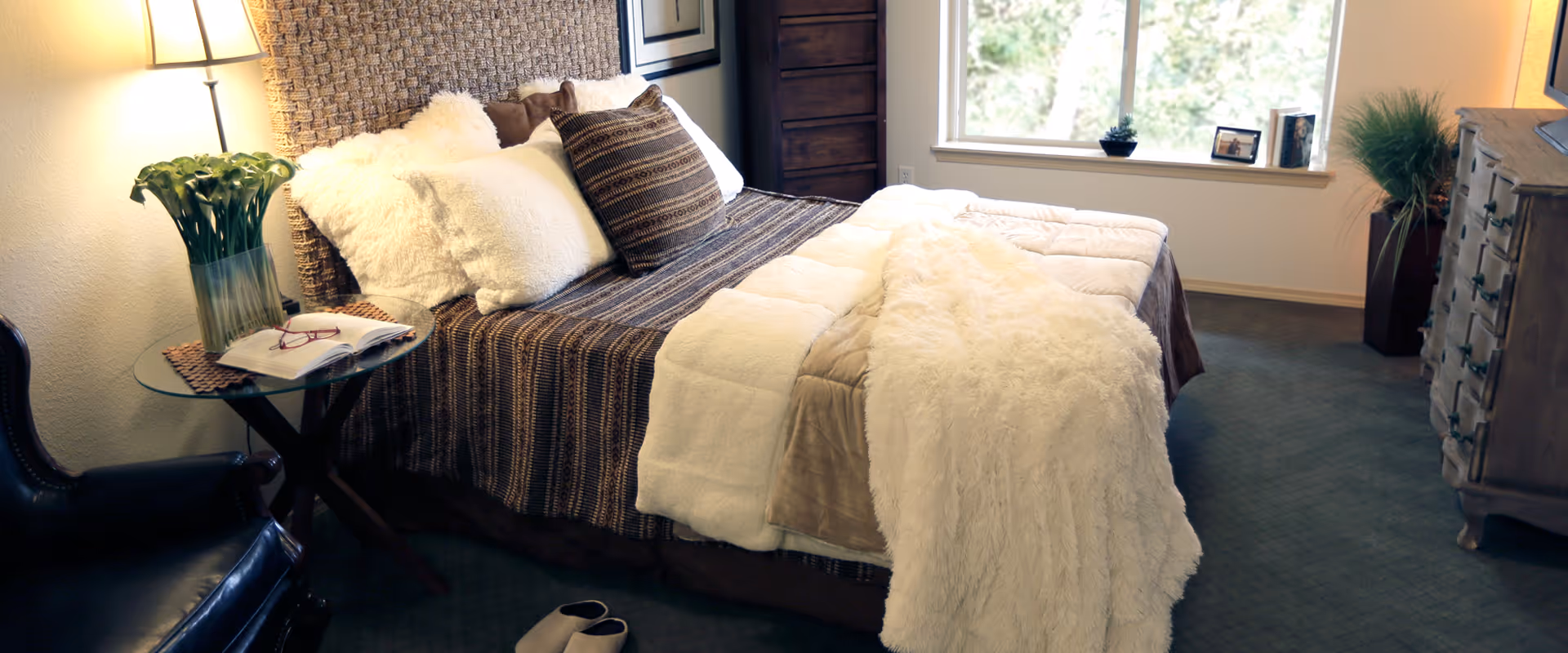 A cozy bedroom with a neatly made bed featuring a brown striped bedspread, white and brown pillows, and a fluffy white throw blanket. A glass-top side table with a vase of flowers and an open book sits next to the bed. There is a leather chair nearby, a wooden dresser, and a window with a view of greenery outside.