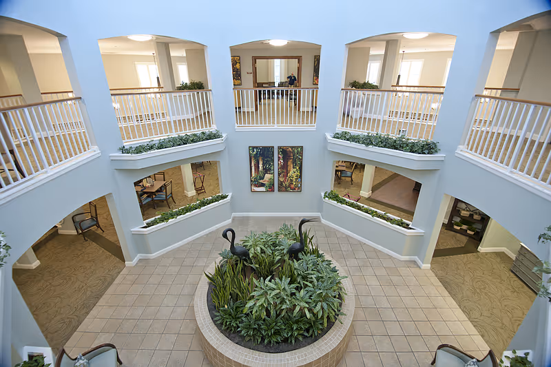 View of a spacious two-story interior common area in a senior living facility with a central circular planter filled with green plants and decorative black bird sculptures. The upper level has white railings and overlooks the lower tiled floor area. There are several seating areas and tables visible on the lower level, with light blue walls and large windows allowing natural light.