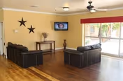 A spacious living room with wooden flooring, two black leather sofas arranged facing each other, a small wooden table with a vase and decorative items against a yellow wall with two black star decorations, a wall-mounted flat screen TV, ceiling fan with light, and large sliding glass doors leading to an outdoor area.