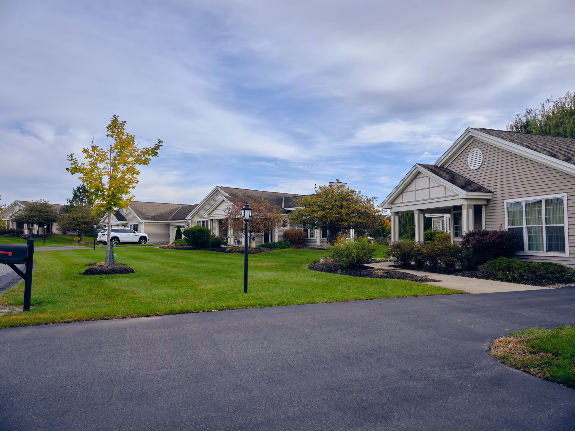 Exterior view of a senior living facility with multiple single-story beige buildings featuring white trim and gabled roofs. The well-maintained lawn has small trees, shrubs, a black mailbox, and a street lamp under a partly cloudy sky.