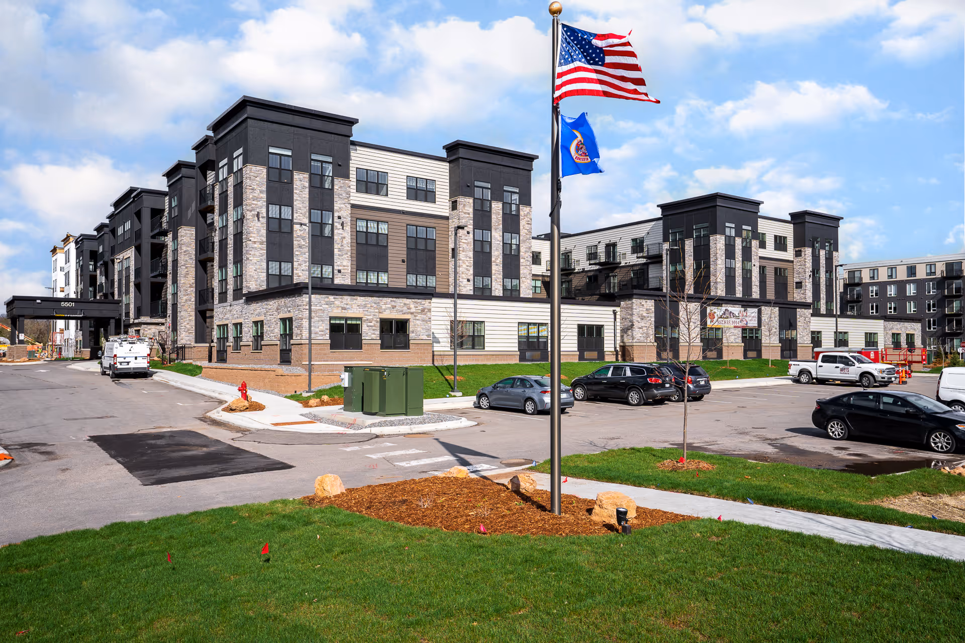 Exterior front view of a multi-story senior living building with American and state flags, parked cars, and landscaped lawn.