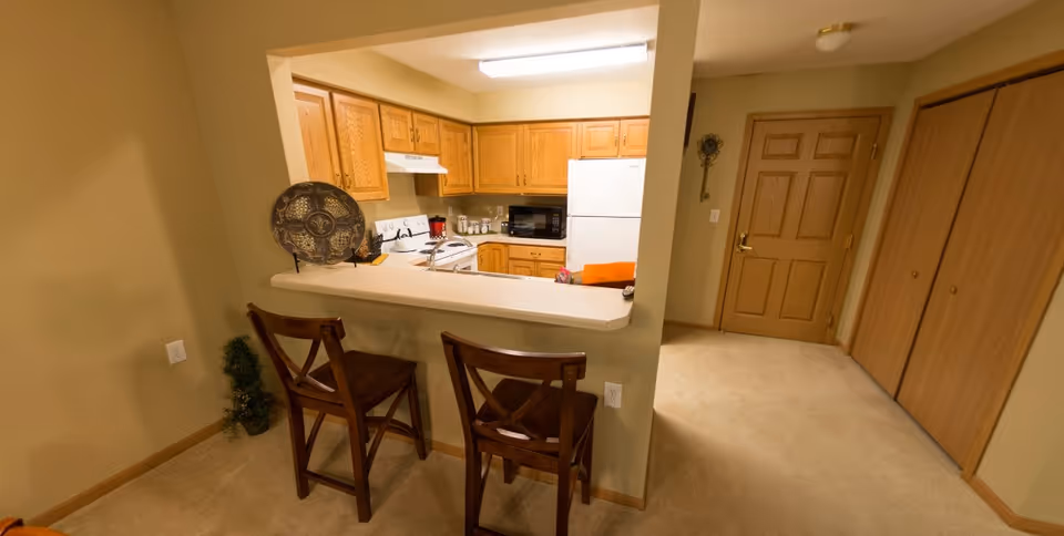 Interior view of a small kitchen area with wooden cabinets, a white refrigerator, stove, and microwave. In front of the kitchen counter are two wooden bar stools. The surrounding area has beige walls and carpeted floors, with a wooden door and closet doors visible in the background.