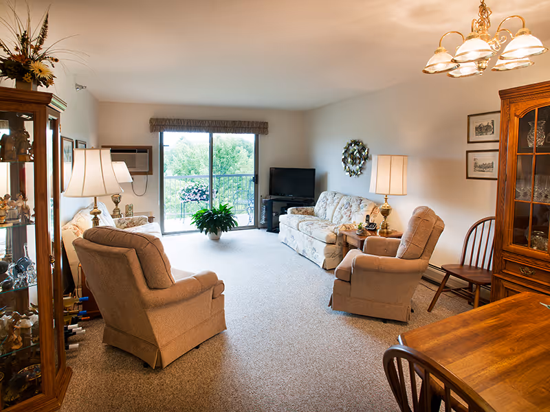 A cozy senior living room with two beige armchairs and a floral sofa arranged around a small wooden table with a lamp. There is a TV on a stand in the corner near a sliding glass door that opens to a balcony with greenery outside. A wooden china cabinet with glassware and decorative items is on the right, and a wooden dining table with chairs is partially visible in the foreground. The room is softly lit with ceiling lights and table lamps.