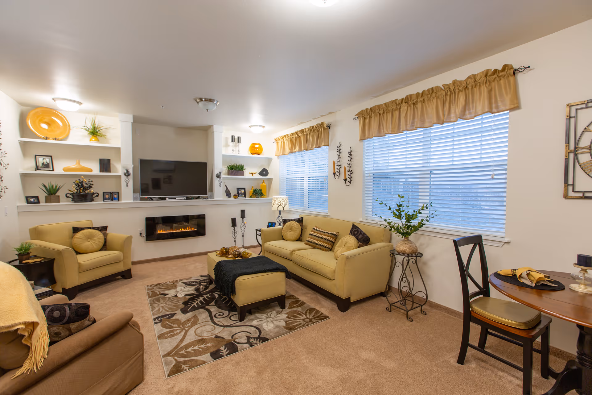 Well-lit living room with beige sofas and armchairs around a patterned rug, a wall-mounted TV above an electric fireplace, decorative shelving, and a small dining table by the windows.