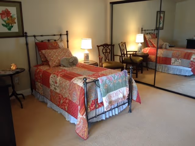 A cozy bedroom with a single iron bed covered in a red and beige patchwork quilt. There is a small stuffed elephant on the bed. Next to the bed is a nightstand with a lit lamp. Two wooden chairs are placed against a mirrored closet door reflecting the room. A framed floral picture hangs on the wall above a small round table with a decorative item on it.